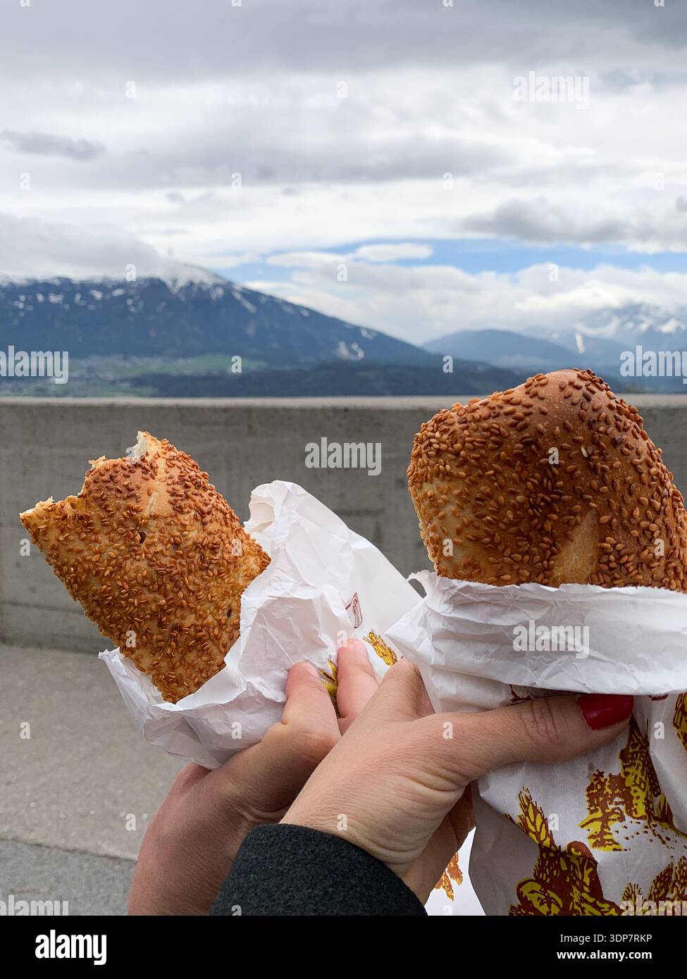 Zwei Touristen, die Sandwiches mit den Bergen im Hintergrund halten. Blick auf Innsbruck im April. Panoramablick auf Innsbruck und Bergkette. - Smartphone-aufgenommenes Stockfoto