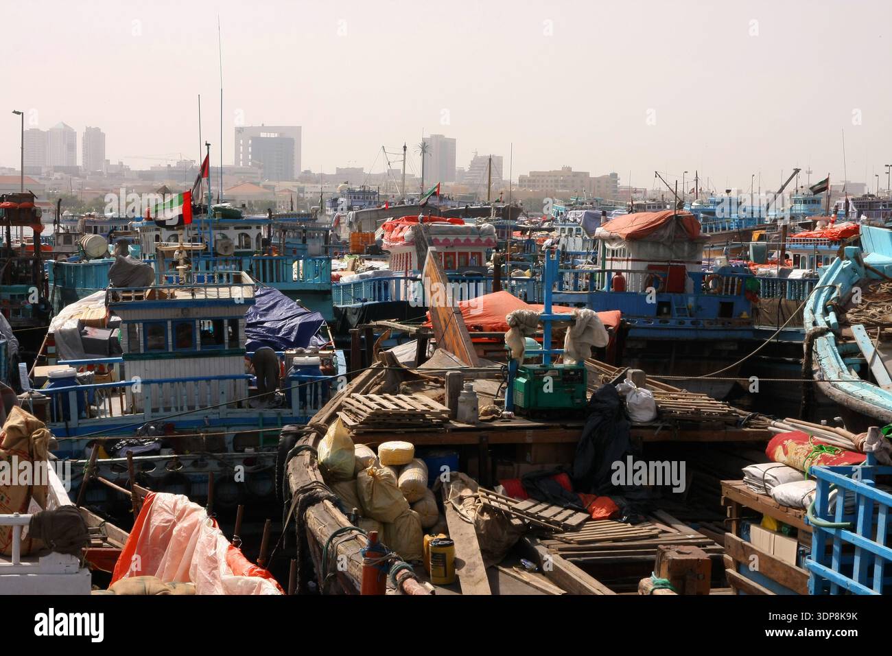 Zahlreiche traditionelle hölzerne Fracht-Dhow-Boote drängen sich an einem belebten Hafen entlang des Dubai Creek mit Stadtgebäuden im Hintergrund. Stockfoto