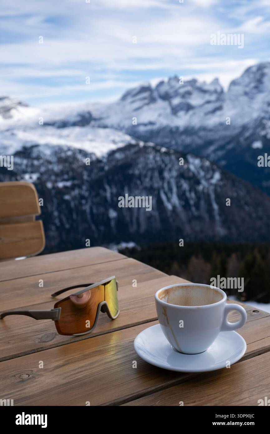 Tasse Kaffee auf einer Holzterrasse mit Sonnenbrille und Panoramablick auf die schneebedeckten Berge, entspannendes Winterreisekonzept Stockfoto
