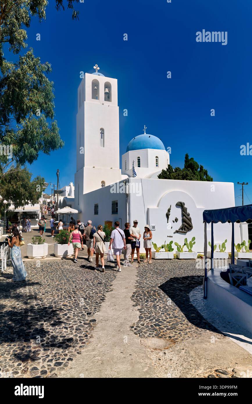 Touristen erkunden eine bezaubernde Kopfsteinpflasterstraße in Santorin, Griechenland, vorbei an berühmten weißen Gebäuden mit blauen Kuppelkirchen unter klarem Himmel. Stockfoto