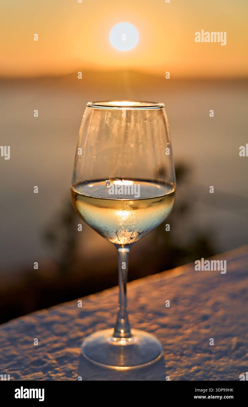 Ein Glas gekühlter Weißwein fängt das goldene Licht eines ruhigen Sonnenuntergangs über dem Meer ein und schafft eine ruhige und malerische Abendszene. Stockfoto