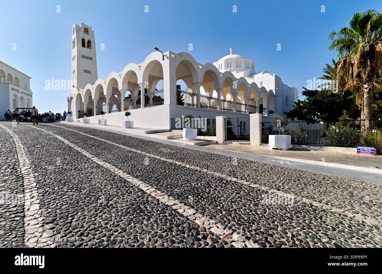 Touristen laufen auf einer Kopfsteinpflasterstraße vorbei an der weißen Metropolitan Cathedral of Thira und dem Hotel Atlantis in Fira, Santorin, Griechenland. Stockfoto