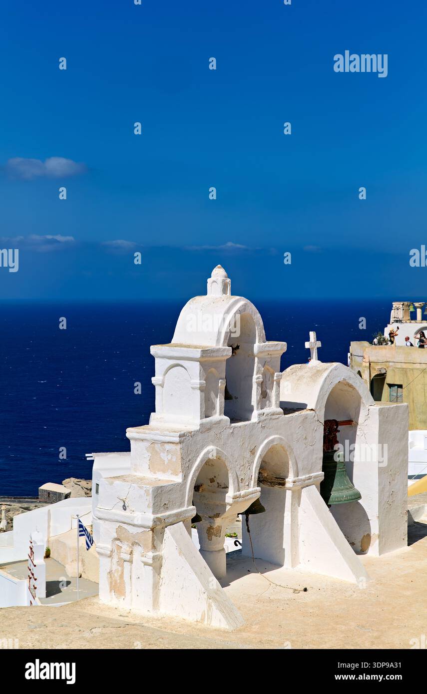 Ein traditioneller weißer Kirchturm überblickt die tiefblaue Ägäis in Santorin, Griechenland. Touristen halten die ikonischen Aussichten der Kykladen fest. Stockfoto