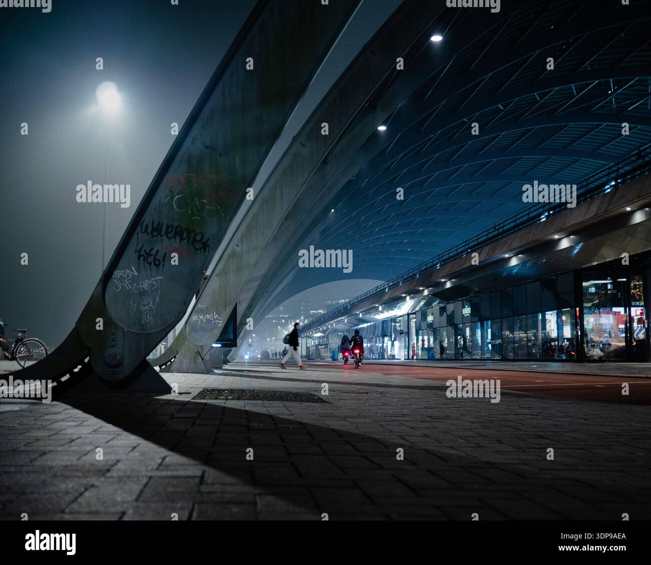 Amsterdam, Niederlande - 13. Februar 2025: Moderne Brückenarchitektur in Amsterdam in einer nebeligen Nacht. Fahrräder und Fußgänger auf beleuchteter Stadtstraße Stockfoto