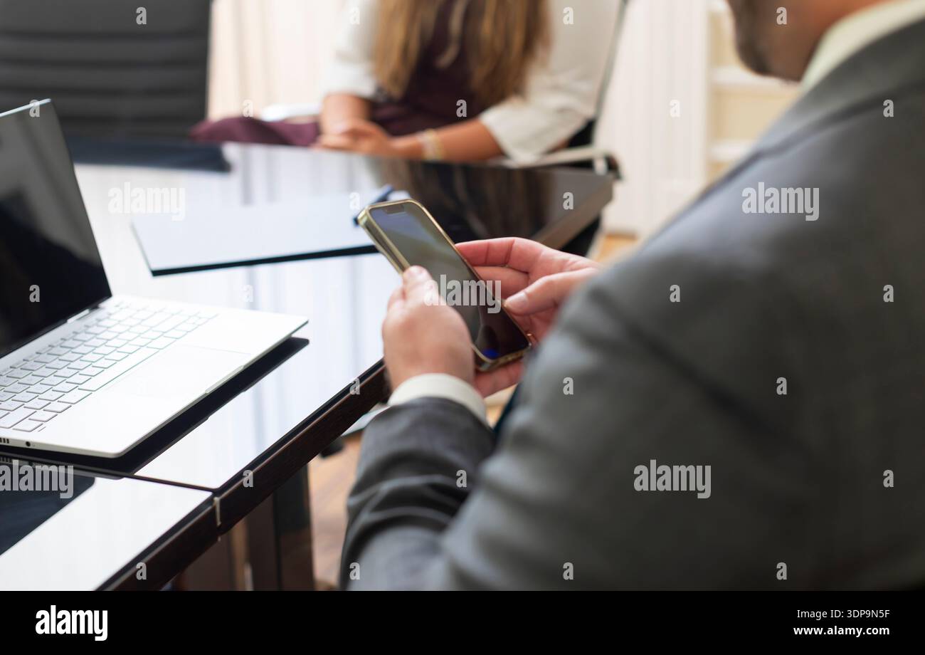 Eine Person im Anzug schaut auf ein Mobiltelefon, während sie an einem schwarzen Tisch sitzt. Eine andere Person sitzt im Hintergrund. Ein Laptop befindet sich auf dem Stockfoto