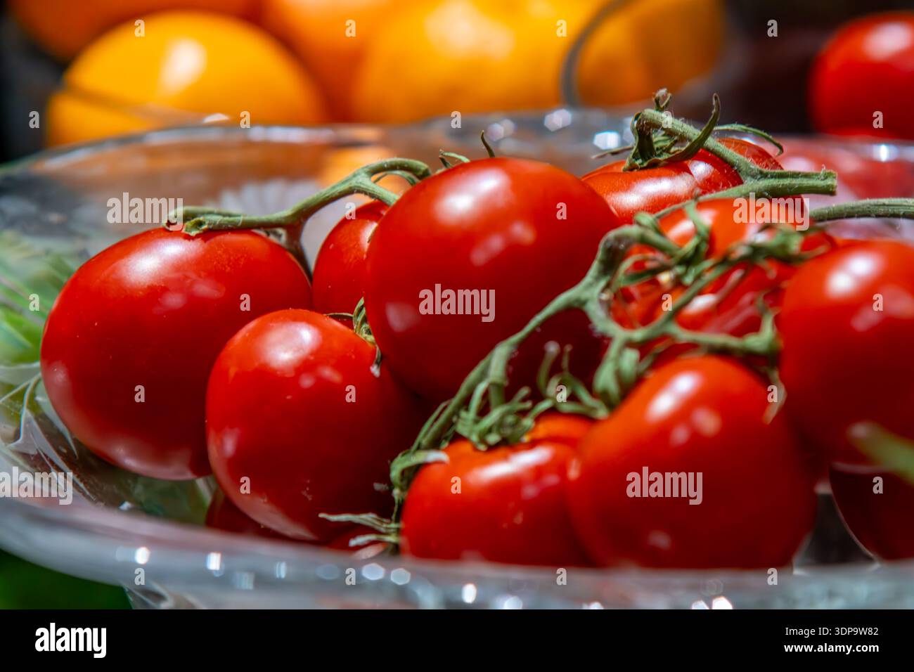 Frische rote Tomaten auf der Rebe in einer Glasschale mit bunten Produkten im Hintergrund. Stockfoto