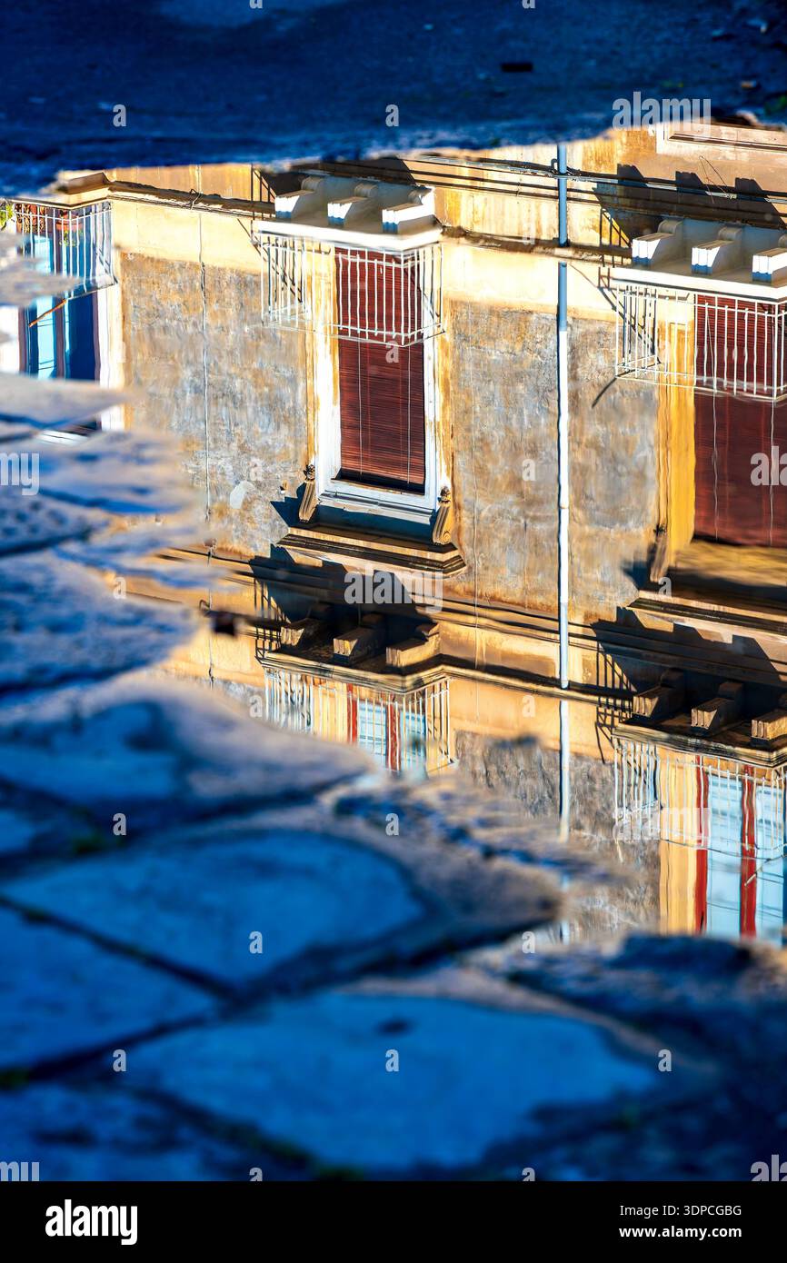 Reflexion von Wohngebäuden mit Balkonen und Fensterläden in einer Wasserpfütze auf Steinpflaster im historischen Zentrum von Catania, Italien. Stockfoto