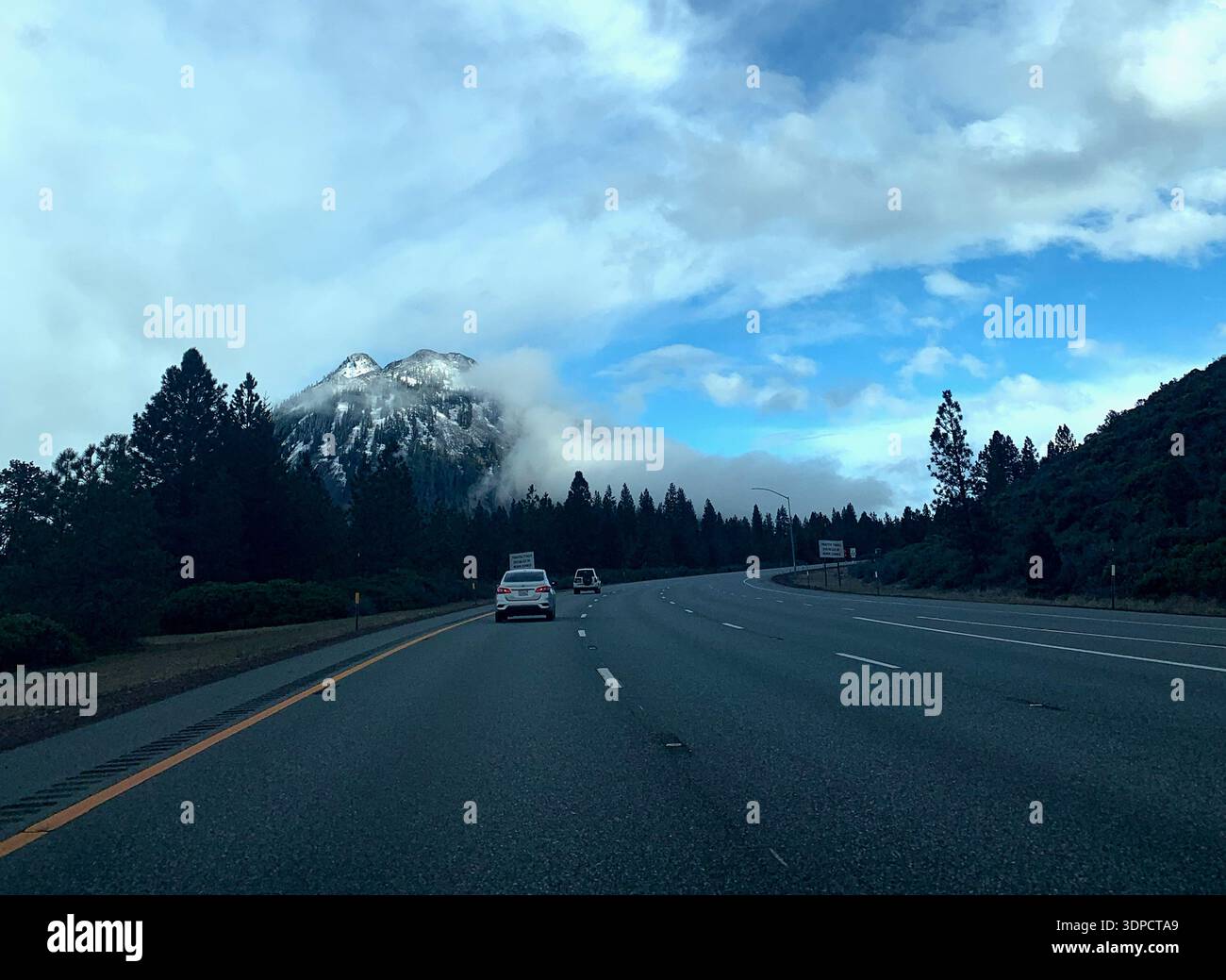 Panoramablick auf die Autobahn mit Fahrzeugen, die in Richtung schneebedeckter Berge unter teilweise bewölktem Himmel fahren, umgeben von immergrünen Bäumen und üppiger Vegetation Stockfoto