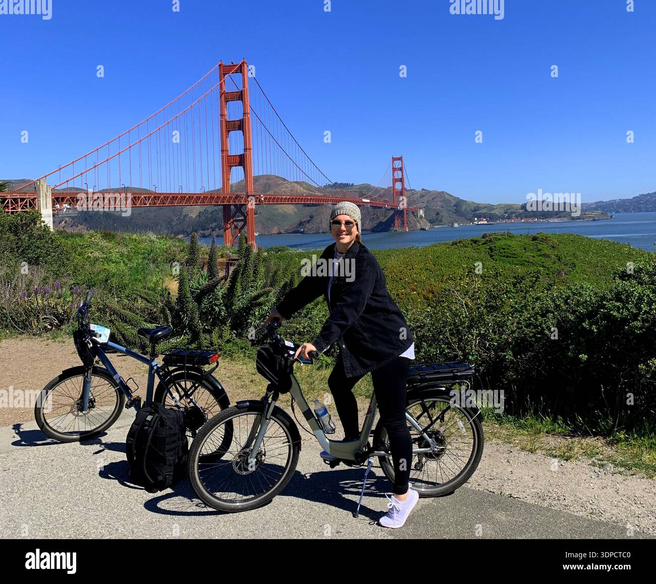 Frau, die mit dem Elektrofahrrad in der Nähe der Golden Gate Bridge mit üppigem Grün und blauem Himmel im Hintergrund unterwegs ist und ein landschaftlich reizvolles Erholungsgebiet im Freien bietet Stockfoto
