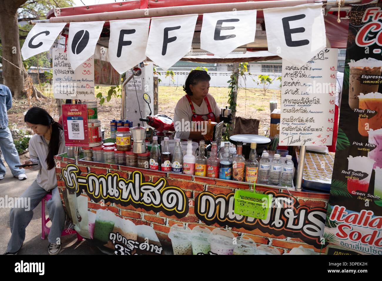 Eine authentische Straßenszene in Thailand, in der eine einheimische Frau Getränke an ihrem farbenfrohen Straßencafé zubereitet. Ein junger Kunde wartet geduldig Stockfoto