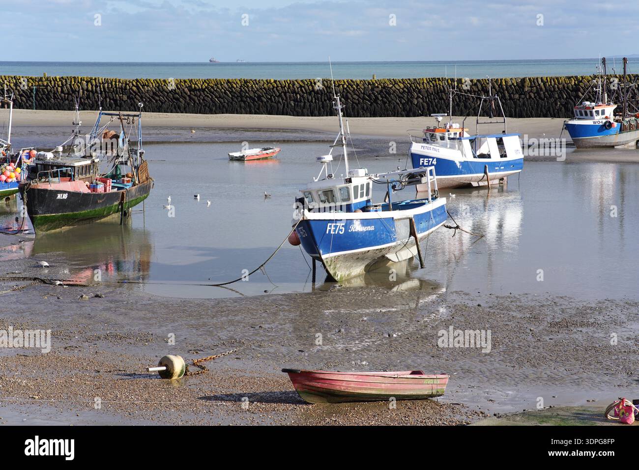 Farbenfrohe Fischerboote bei Ebbe am schlammigen Folkestone Beach, blaue Rümpfe und Reflexionen, Wellenbrecher und ruhiges Meer, Kent England Küste. Stockfoto