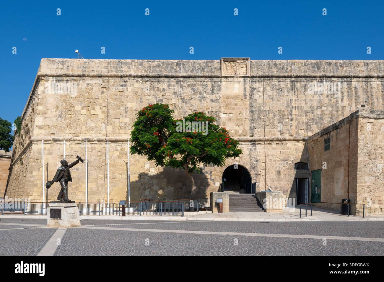 Weiter Blick auf die Statue des Manwel Dimech Monuments auf einem sonnendurchfluteten Valletta-Platz neben massiven Festungsmauern aus Kalkstein und einem bogenförmigen Eingang unter einem Stockfoto