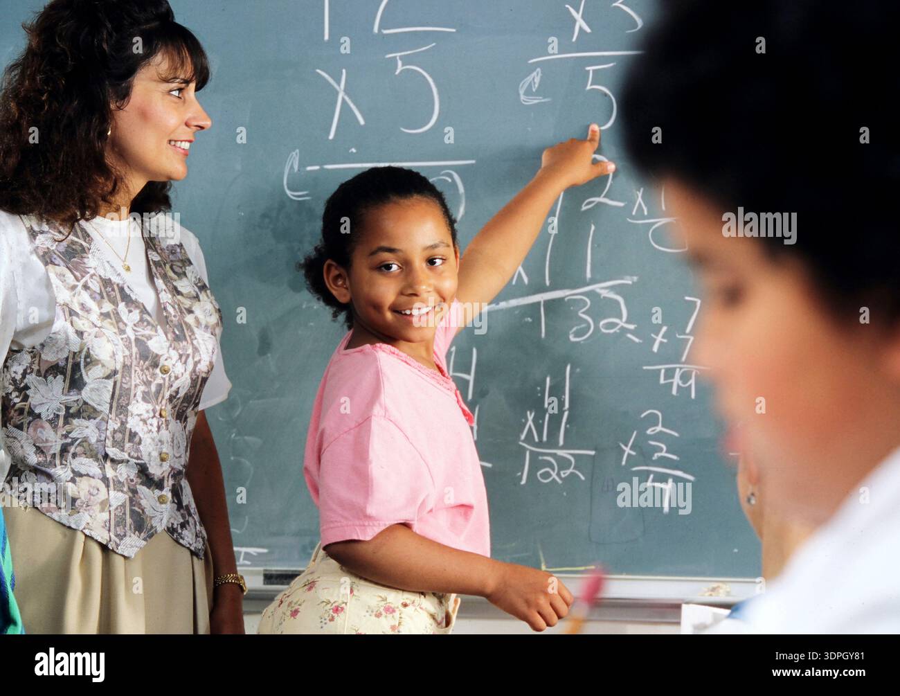 Eine Lehrerin steht zur Seite, als ein Mädchen stolz auf ein Matheproblem auf dem Brett verweist, das sie gelöst hat. Das Klassenzimmer hat ein schwarzes Brett voller Zahlen. Ot Stockfoto