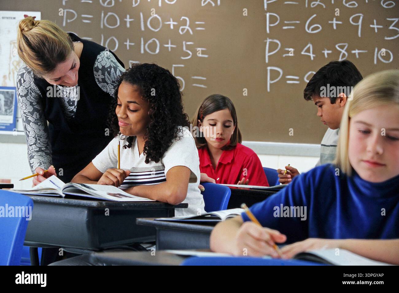 Kinder sitzen an Schreibtischen in einem Klassenzimmer, während ein Lehrer einem afroamerikanischen Schüler mit mathematischen Problemen hilft. Die Lektion konzentriert sich auf Addition und Subtraktion Stockfoto