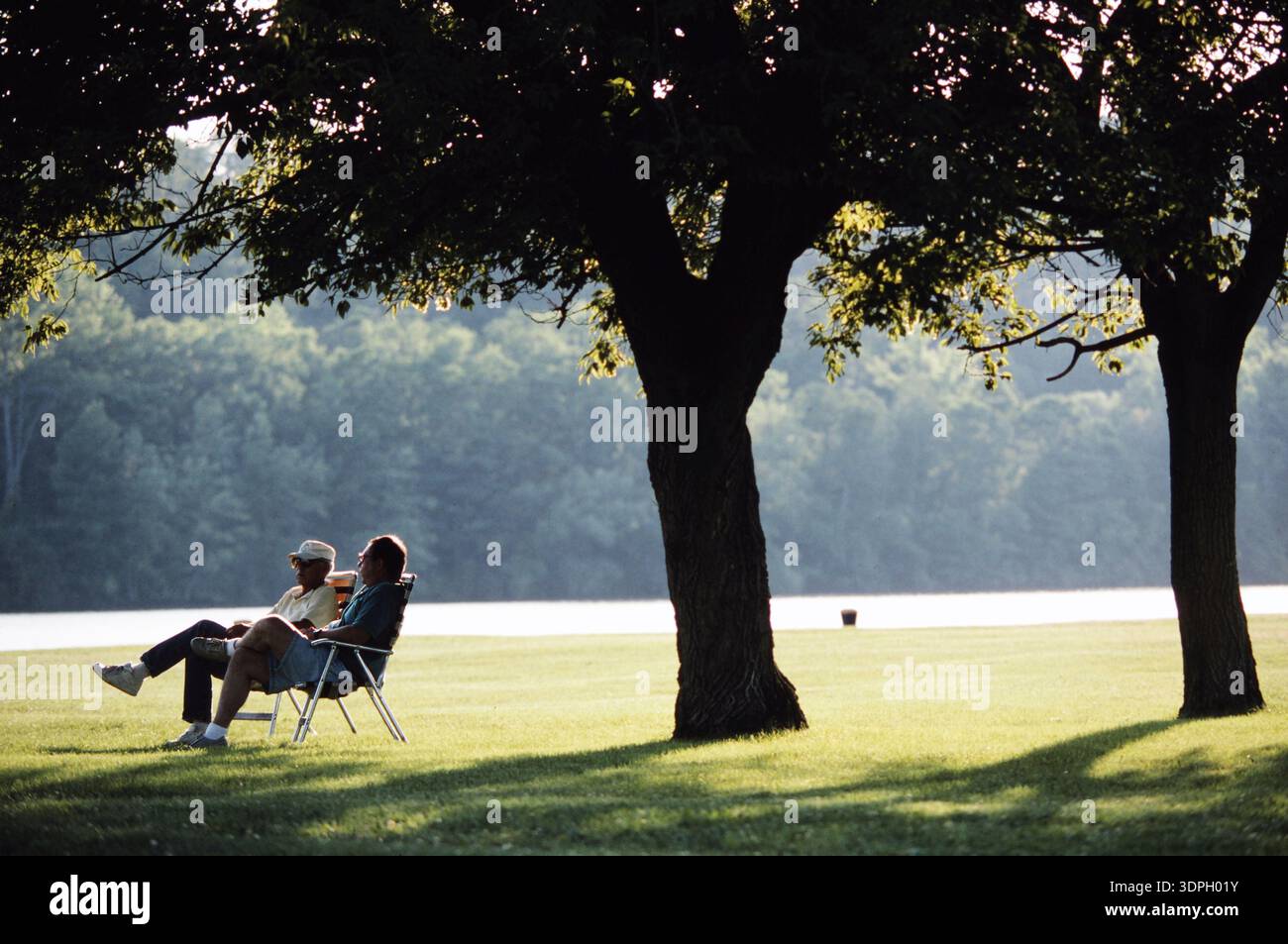 Für pensionierte Männer sitzen auf Liegestühlen unter Bäumen in der Nähe eines Sees. Sie genießen das Sonnenlicht und die friedliche Umgebung. Das Grün und das Wasser bilden einen Quie Stockfoto