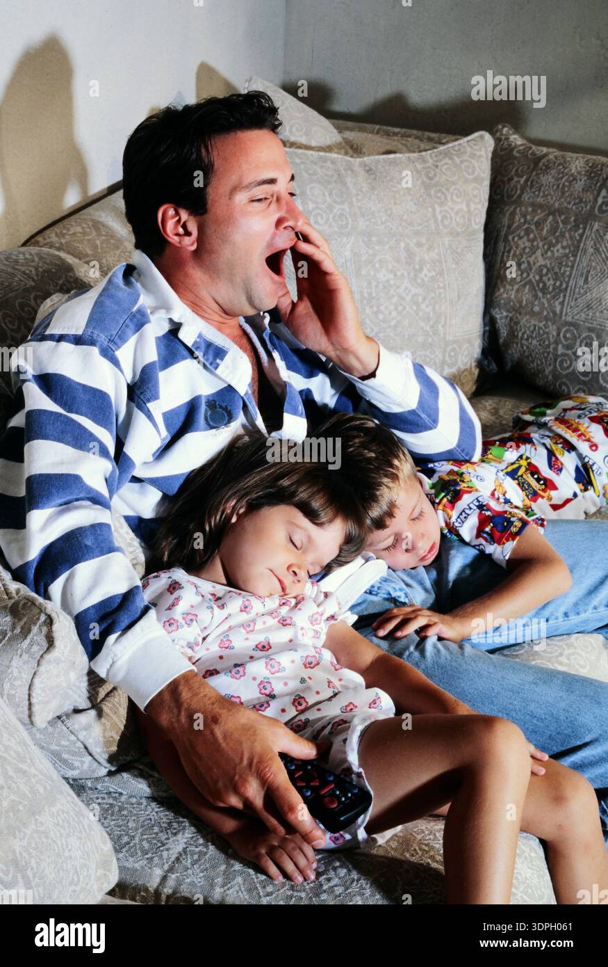 Ein gähnender, verschlafener, erschöpfter Vater sitzt mit seinen beiden Kindern auf einer Couch. Ein Kind schläft auf seinem Schoß, während das andere neben ihm ruht. Der Vater Ya Stockfoto