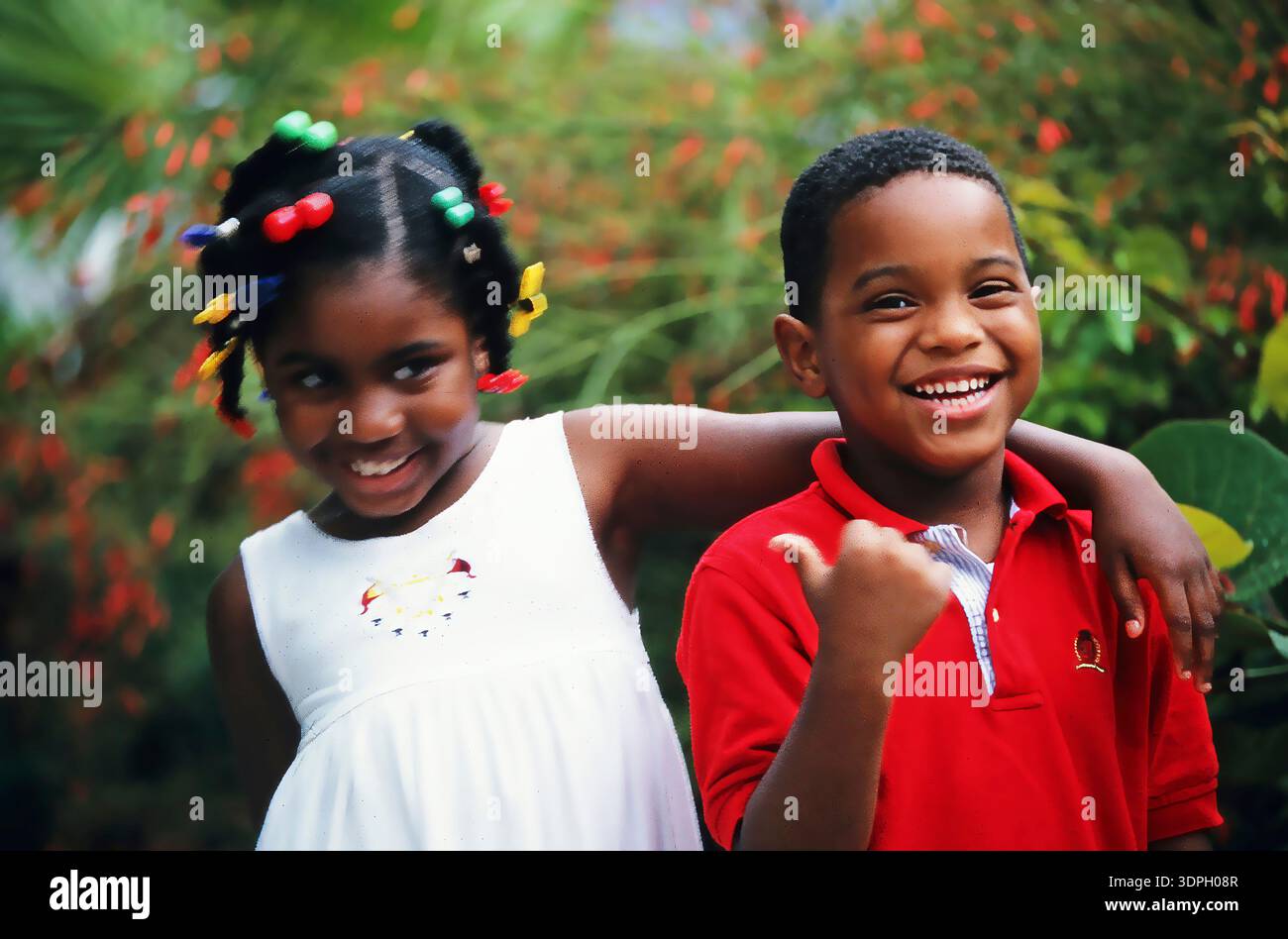 Bruder und Schwester stehen dicht beieinander in einem Garten. Ein Mädchen trägt ein weißes Kleid und schaut seitlich, während ein Junge in Rot lächelt und die Daumen hoch zeigt. Br Garten Stockfoto