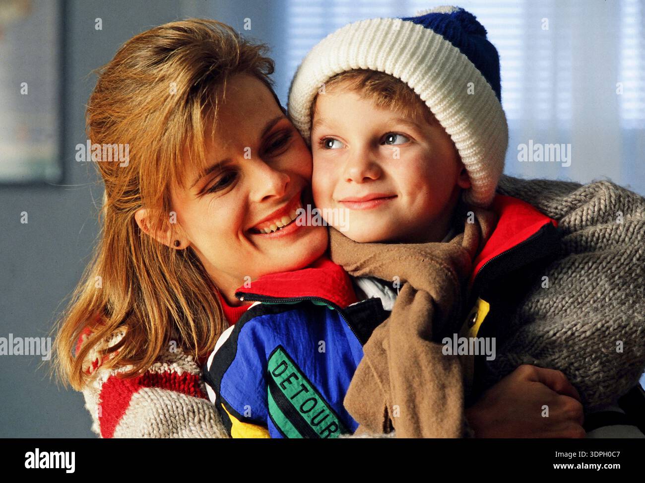 Eine Mutter umarmt ihren Sohn, beide lächeln, in einer warmen Innenumgebung. Der Junge trägt einen Winterhut und einen farbenfrohen Mantel, während die Frau fröhlich aussieht Stockfoto