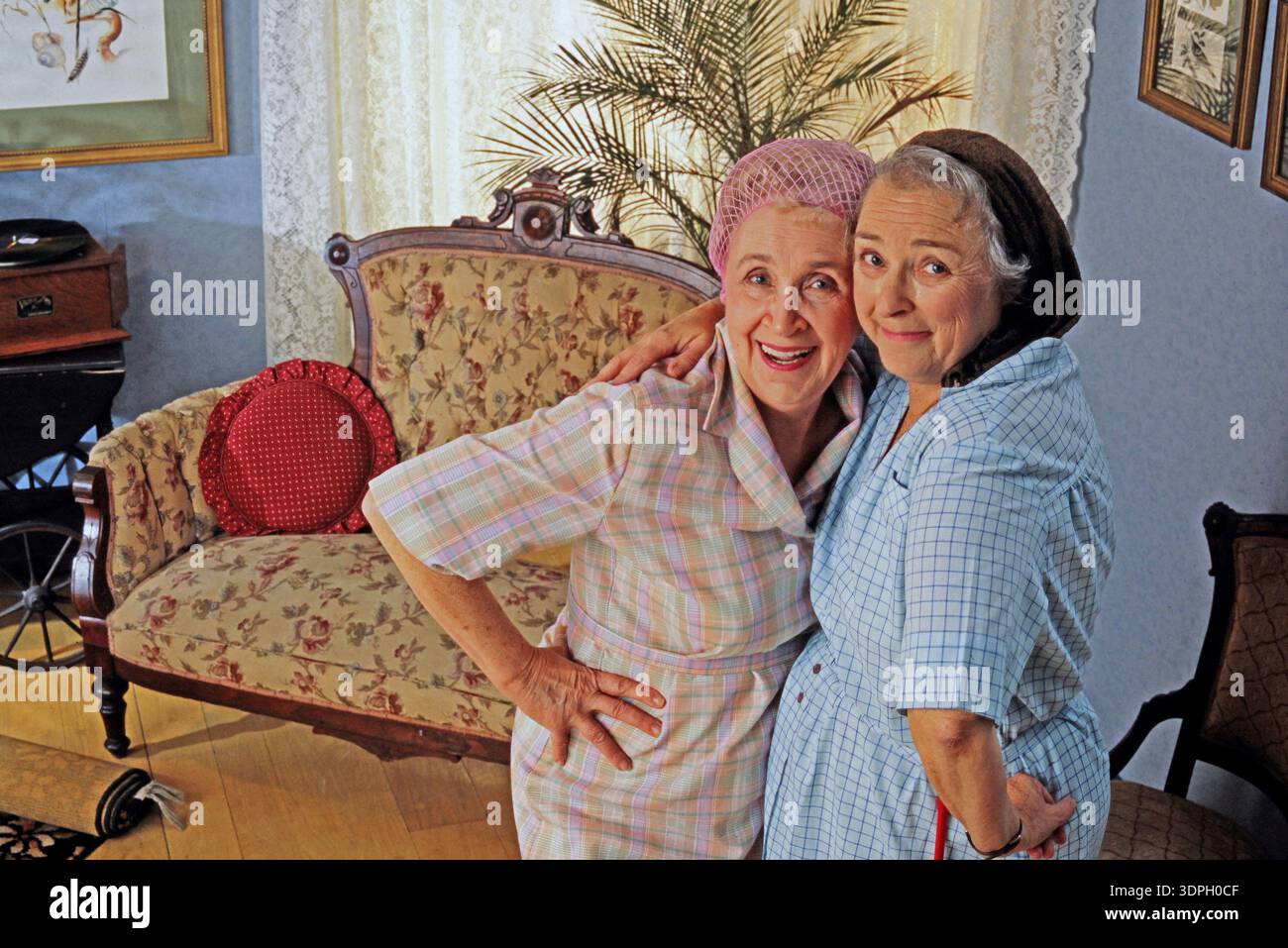 Zwei ältere Freundinnen stehen nahe beieinander und lächeln in einem Vintage-Wohnzimmer. Sie tragen helle Kleidung und Enjo Stockfoto