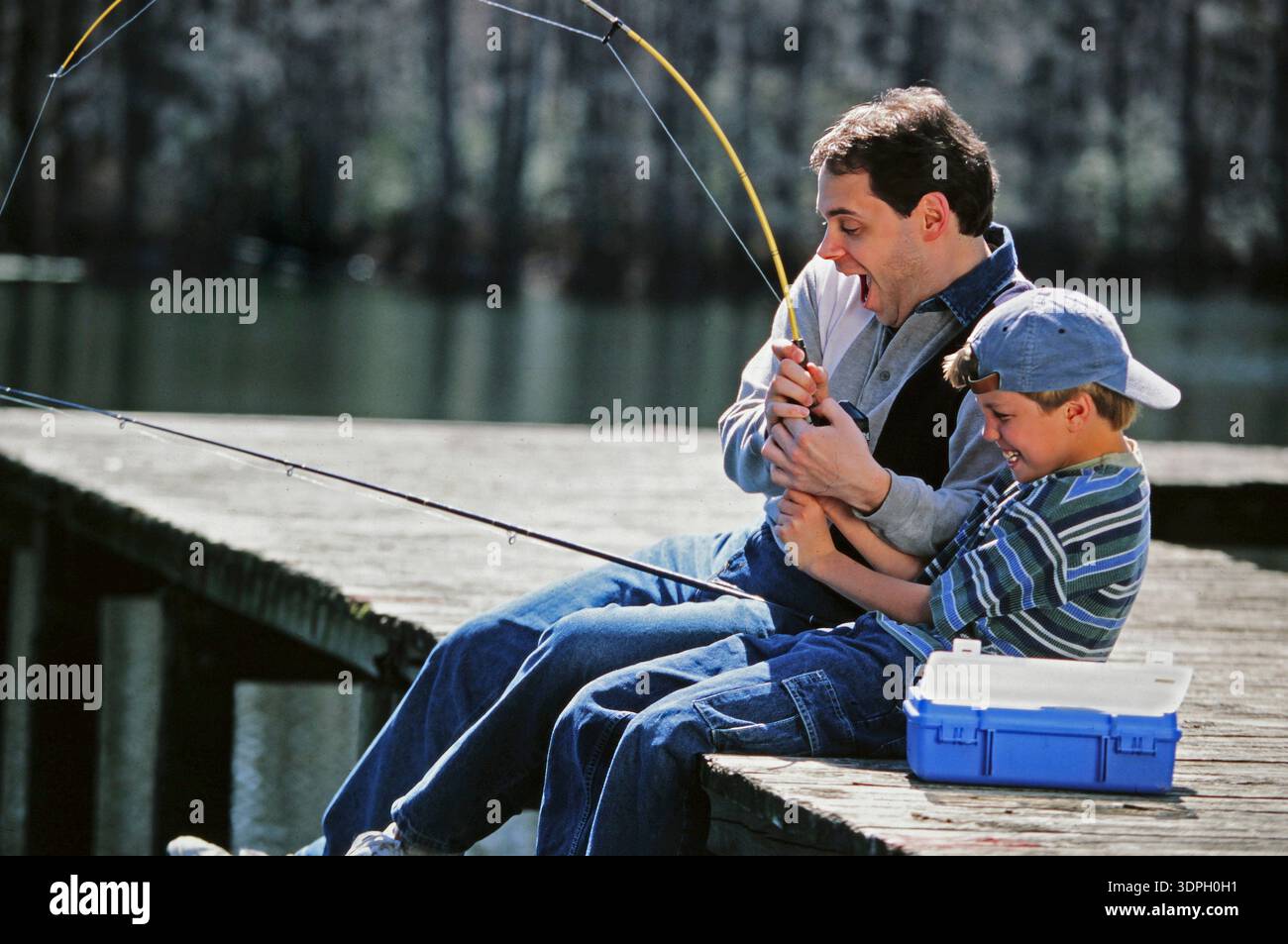 Vater und Sohn sitzen auf einem hölzernen Dock und arbeiten zusammen, um einen Fisch einzurollen. Beide zeigen Aufregung und Freude, während sie ihren Angelausflug am lak genießen Stockfoto
