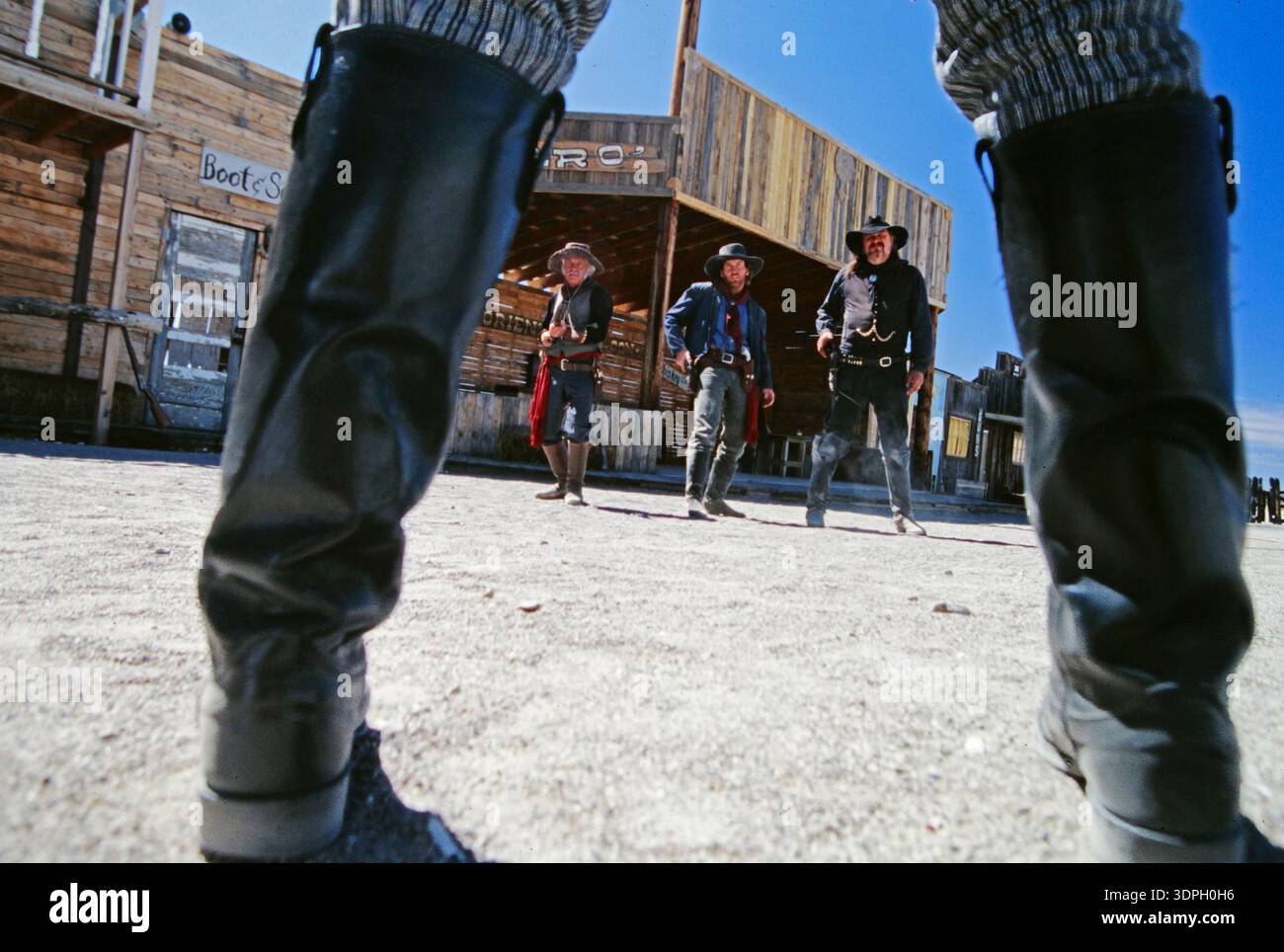 Drei Cowboys stehen in der Geisterstadt TGoumbstone, Arizona, unter einem blauen Himmel, während ein Cowboy seine Stiefel fokussiert Stockfoto