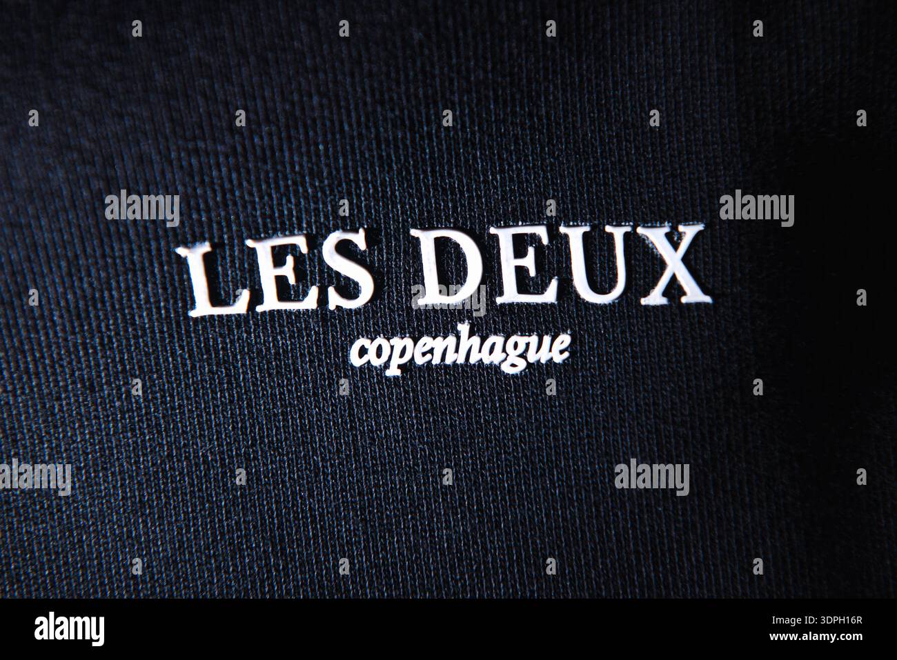 Les Deux Copenhague Logo der Modemarke auf schwarzer Kleidung mit Streulichtschranke. Weißer, erhabener Schriftzug auf dunkler gerippter Stoffstruktur mit künstlerischem lichteinfall Stockfoto