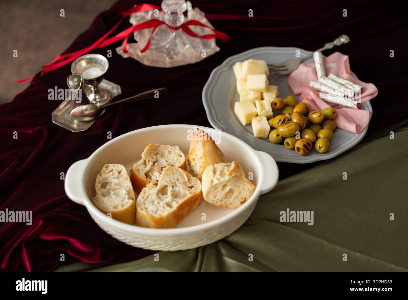 Stilvolle Tischplatte mit geschnittenem Baguette in einer weißen Keramikschale auf einem burgunderroten Samttuch und olivgrün drapierten Tuch. Daneben ist ein Antipa Stockfoto
