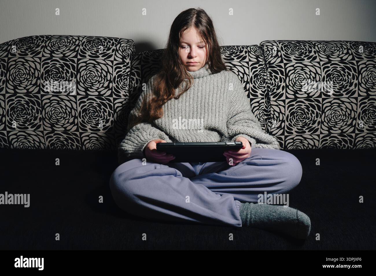 Teenager in warmem Pullover auf dem Sofa sitzen und nachts ein Tablet benutzen. Konzept der Hausunterweisung oder Unterhaltung bei Stromausfall Stockfoto