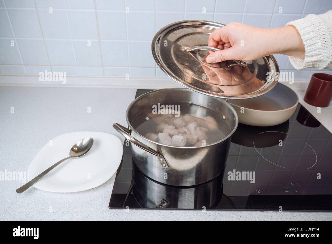 Öffnen Sie einen Topfdeckel aus Edelstahl von Hand, um kochende Fleischbrühe für traditionelle ukrainische Borschtsuppe auf einem modernen Induktionsofen zu prüfen Stockfoto