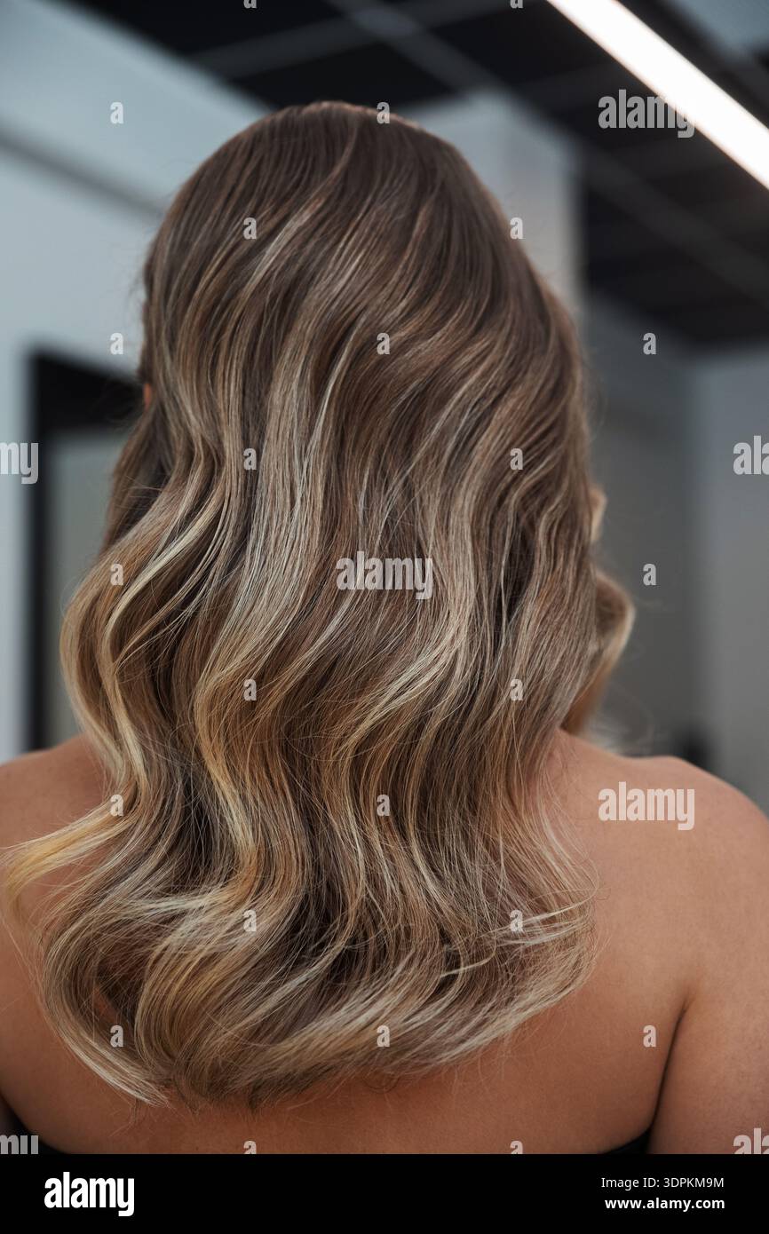 Eine Person mit langen welligen Haaren in Braun- und Blondtönen befindet sich in einem stilvollen Salon. Die Beleuchtung hebt die Struktur und den Fluss des Haares in einem Ko hervor Stockfoto