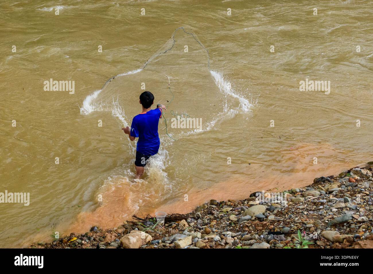Vang Vieng, Laos – 16. Juni 2020: Eine Person steht in seichten Gewässern nahe dem felsigen Ufer des Nam Song River und wirft ein Fischernetz in den schlammigen Fluss. Stockfoto