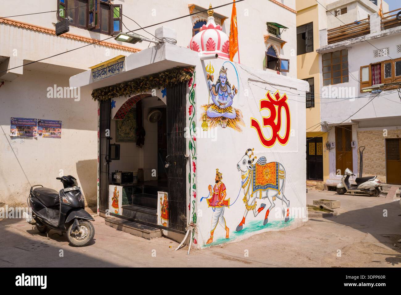 Udaipur, Indien - 23. März 2022: Ein kleiner Hindutempel steht an einer Straßenecke mit Wandgemälden, die Lord Shiva, eine verzierte Kuh, eine Figur mit einem Dreizack und ein großes rotes OM-Symbol auf der weißen Außenwand darstellen, mit in der Nähe geparkten Motorrollern und Wohngebäuden im Hintergrund. Stockfoto