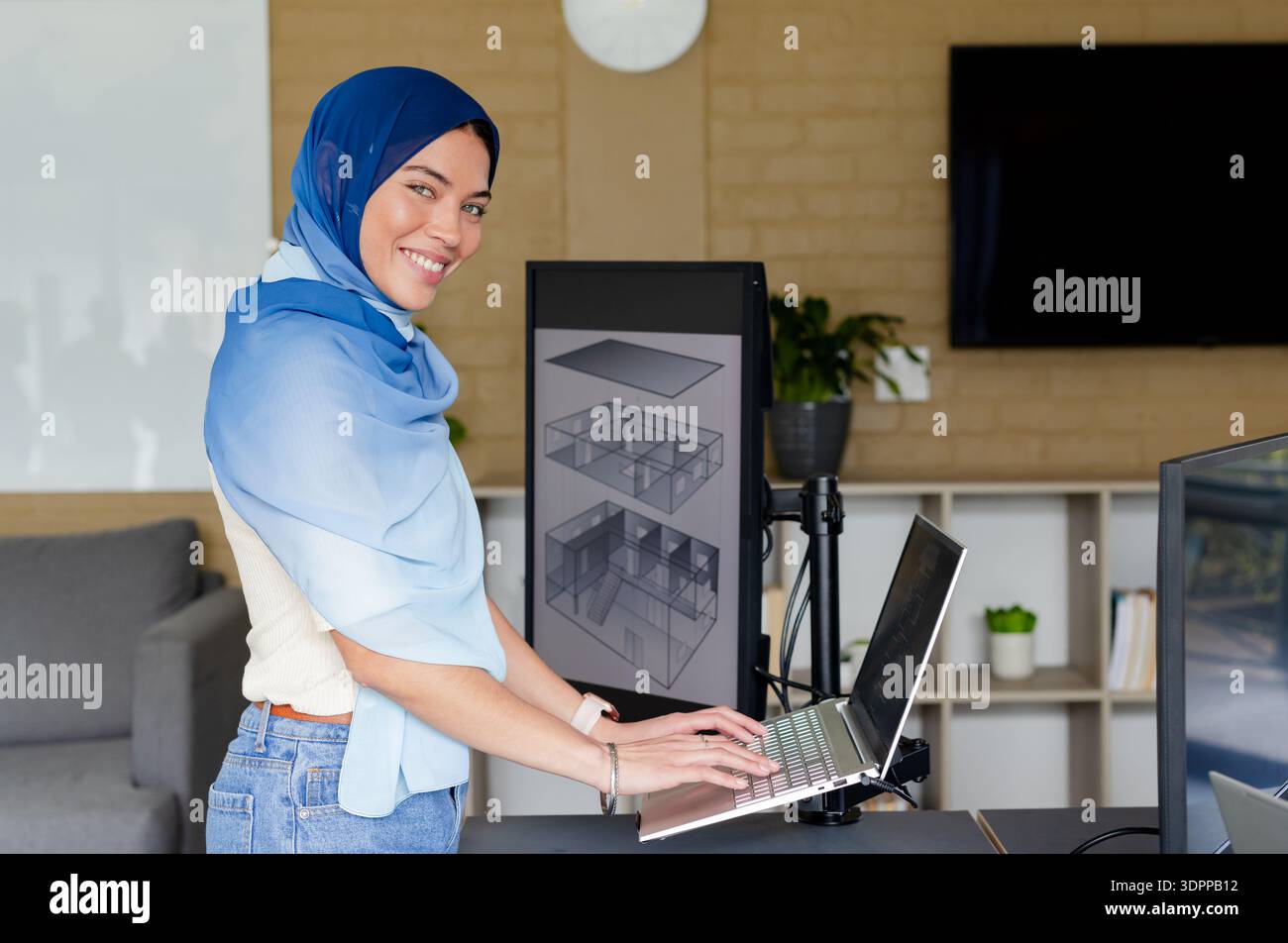 Indische Erwachsene Frau, die auf einem Laptop am Stehpult im Großraumbüro mit 3D-Architekturmodellen tippt Stockfoto