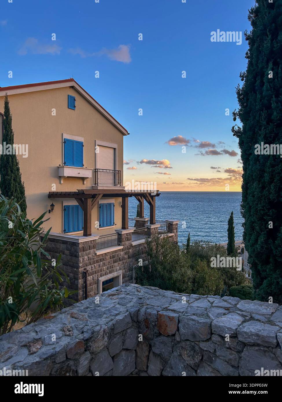 Charmante mediterrane Villa mit blauen Fensterläden und Blick auf das Meer bei Sonnenuntergang, eingerahmt von Zypressen. - Smartphone-aufgenommenes Stockfoto