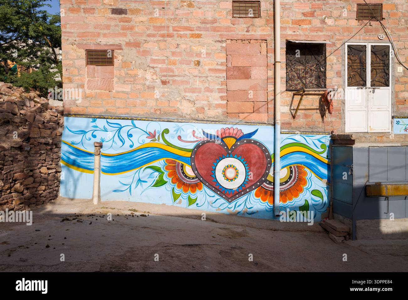 Jodhpur, Indien - 26. März 2022: An der unteren Wand eines Wohngebäudes aus Backstein ist ein lebendiges Wandbild mit einem großen Herzmotiv mit Blumen- und Wellenmustern gemalt, mit einer weißen Tür, Gitterfenstern und sichtbaren Steinmetzwerken. Stockfoto