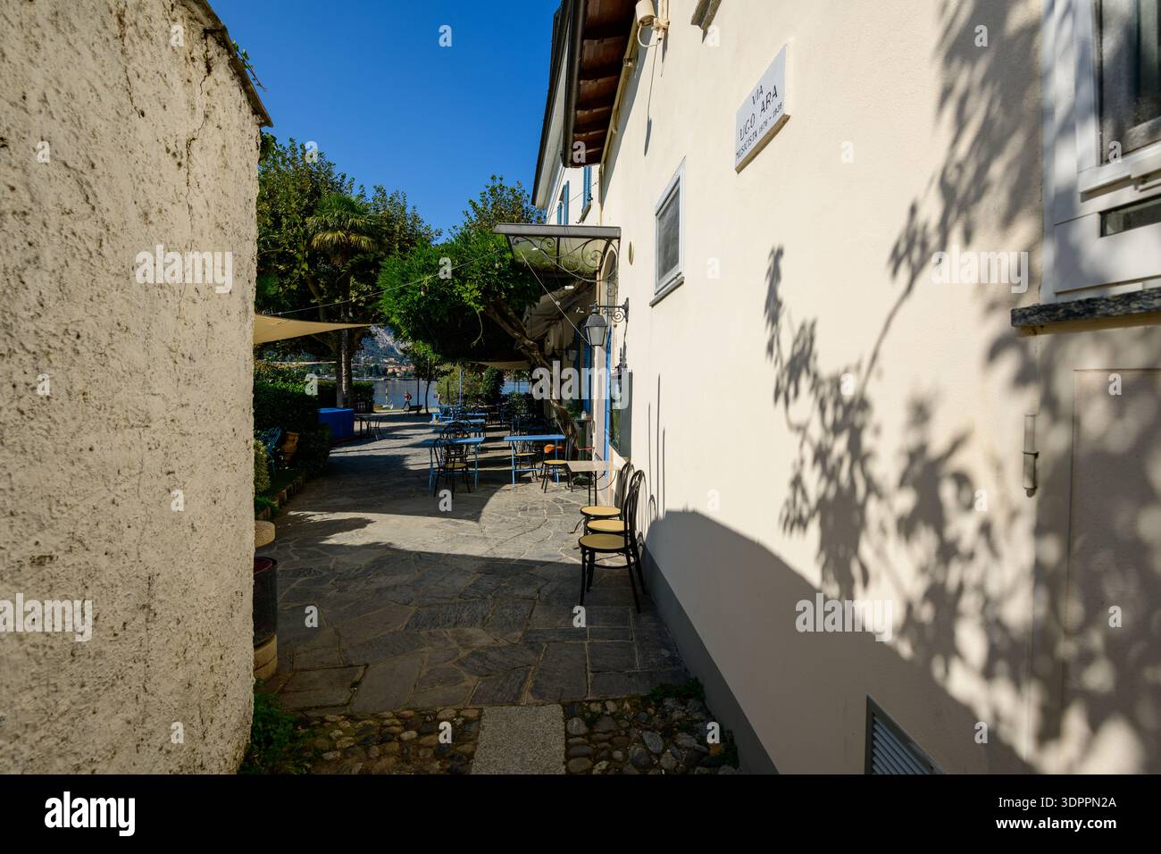 Isola dei Pescatori, Italien - 30. September 2020: Eine schmale Steingasse führt zu einem Café-Sitzbereich im Freien mit Metalltischen und -Stühlen, beschattet von Bäumen und Markisen, mit einem Straßenschild zur Via Lungo Lago an der Gebäudewand. Stockfoto