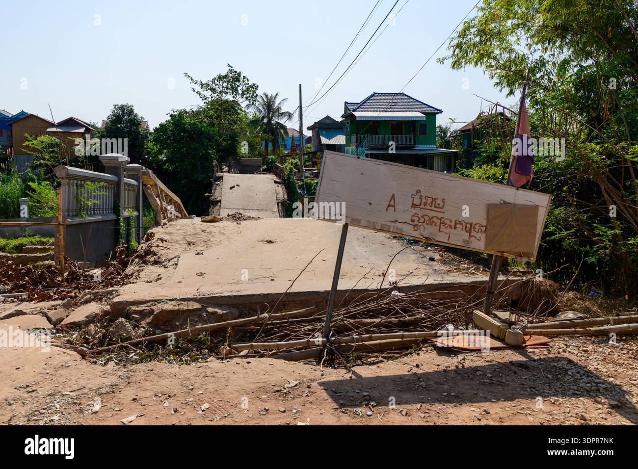 Kambodscha - 7. Februar 2020: Eine beschädigte Betonbrücke ist teilweise eingestürzt und durch ein geneigtes hölzernes Warnschild mit Khmer-Schriftzug blockiert, wobei Häuser, Vegetation und Versorgungsdrähte im Hintergrund unter klarem Himmel sichtbar sind. Stockfoto