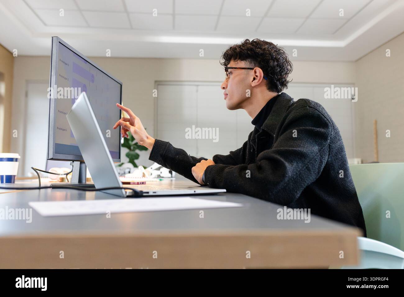 Asiatischer Mann mit Brille, der Daten auf dem Monitor analysiert, Laptop neben Kaffeetasse im Büro, Kopierraum Stockfoto