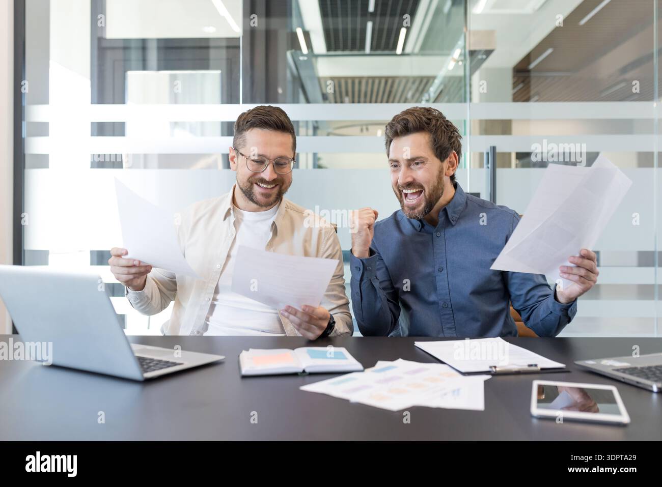 Zwei glückliche männliche Geschäftspartner feiern gemeinsam im modernen Büro Erfolge und finanziellen Erfolg und sehen sich die Dokumente mit Spannung und Erfolg an Stockfoto