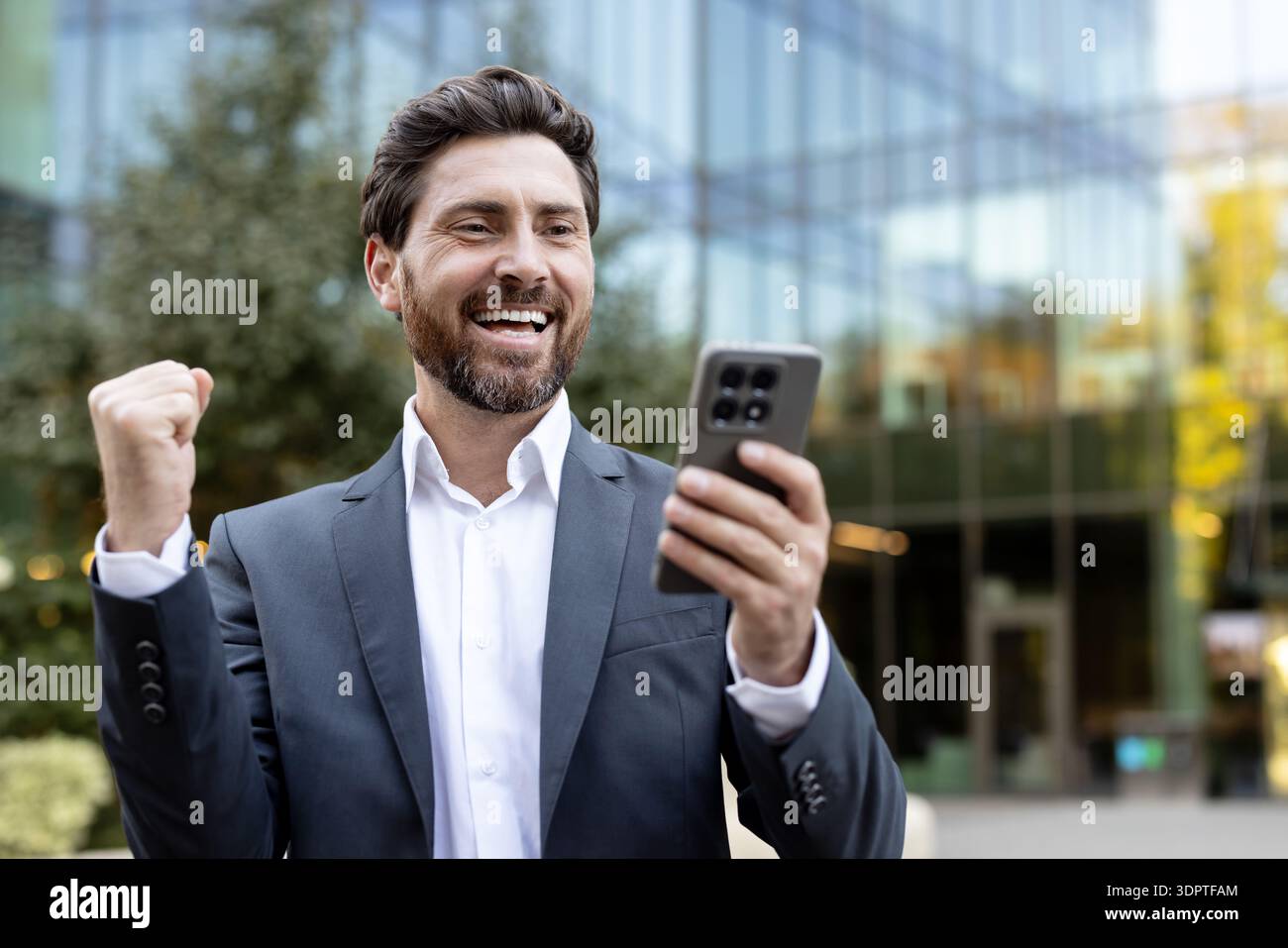 Glücklicher junger bärtiger Geschäftsmann in formeller Kleidung, der draußen steht. Feiert eine Triumphgeste, während er sein Smartphone mit einem großen Lächeln betrachtet. Repräsentiert Online-Erfolg und positive Neuigkeiten Stockfoto