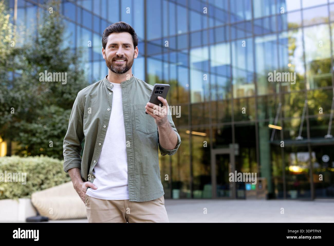 Ein lächelnder Mann, der ein Smartphone hält und selbstbewusst mit einer Hand in der Tasche vor einem modernen Glasbürogebäude steht, repräsentiert urbane professionelle Kommunikation und digitalen Lebensstil Stockfoto