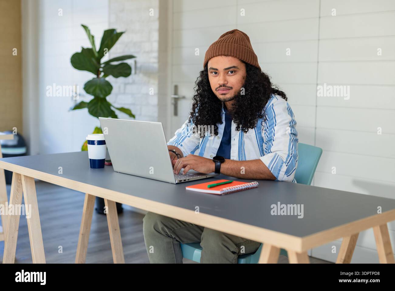 Mann, der am Schreibtisch im Heimbüro mit wiederverwendbarer Kaffeetasse, Notizbuch und farbigen Stiften tippt Stockfoto