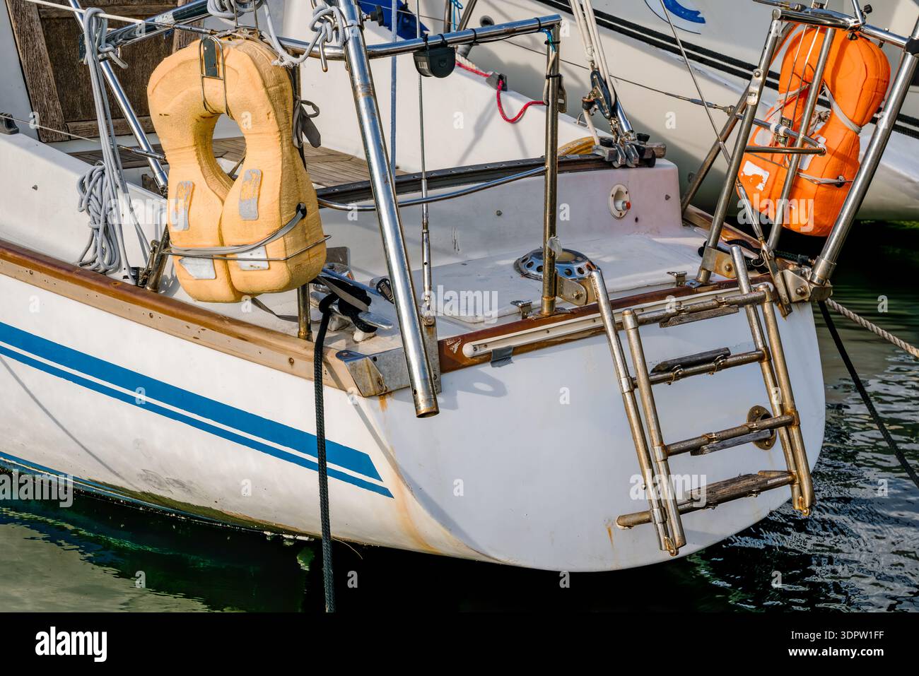 Eine Nahansicht des Hecksterns eines Segelbootes mit einer Metallleiter und zwei Schwimmwesten in Orange und Gelb. Das Boot liegt in ruhigem Wasser. Stockfoto