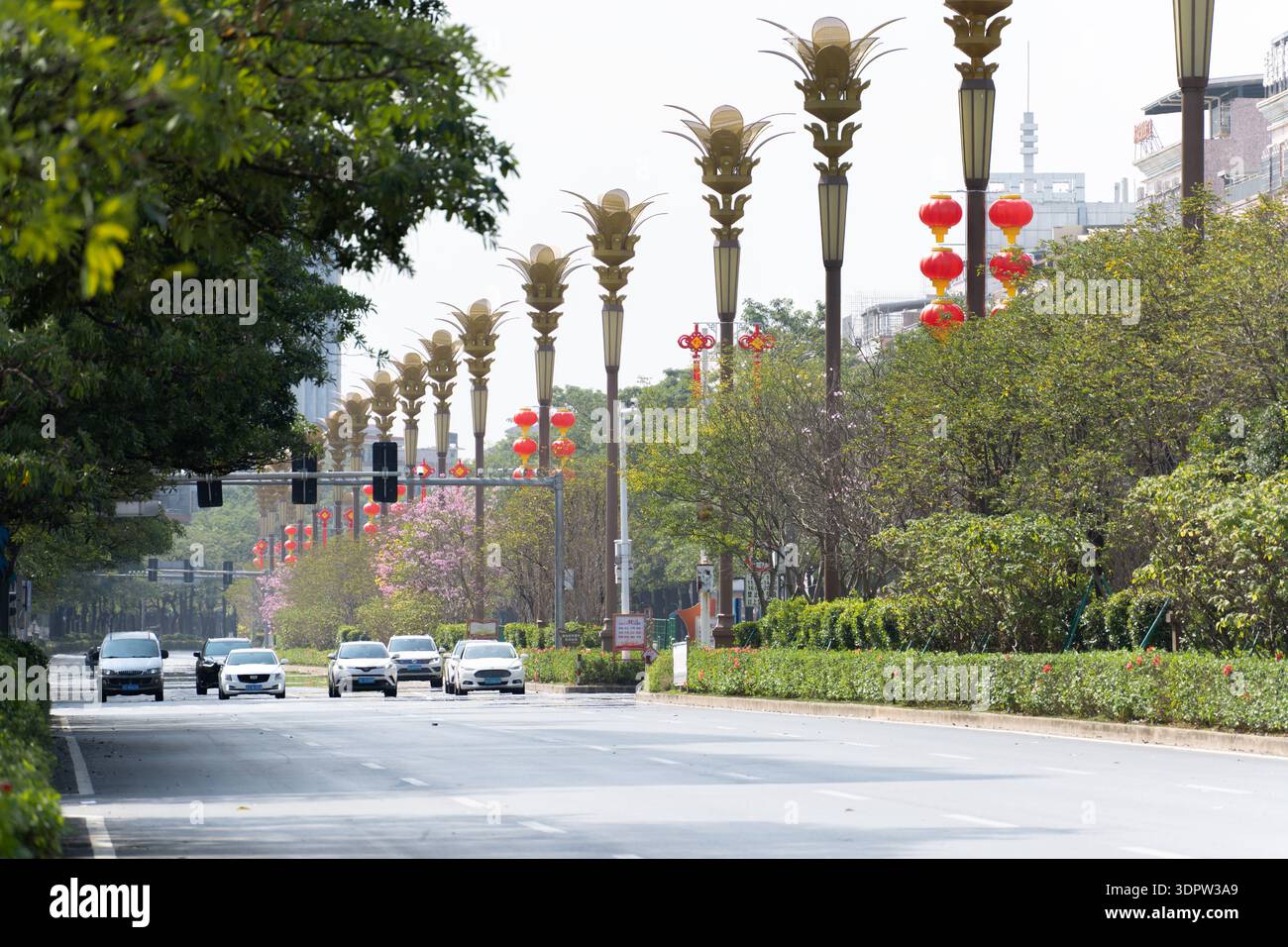 Breite Stadtstraße in China mit palmenförmigen Ampeln, roten Laternen, grünen Bäumen und Autos, die unter bewölktem Himmel fahren Stockfoto