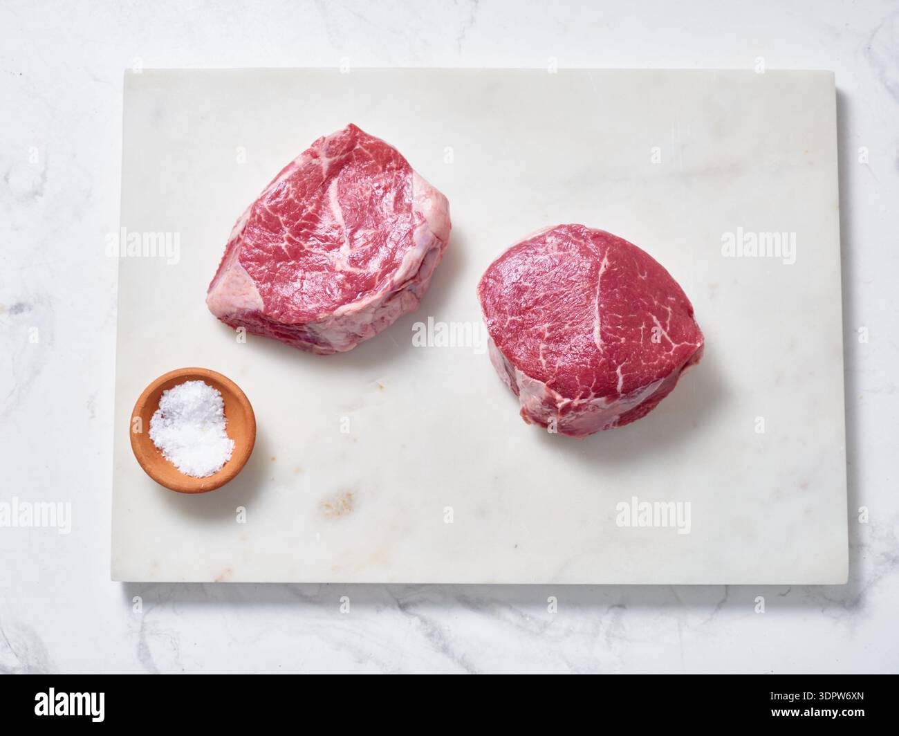 Rohe Chuck zartes Steak auf einem Schneidebrett aus Marmor mit einer kleinen Schüssel Salz daneben. Frische Kräuter und Pfeffer sind in der Nähe. Für din zubereitete Lebensmittel Stockfoto