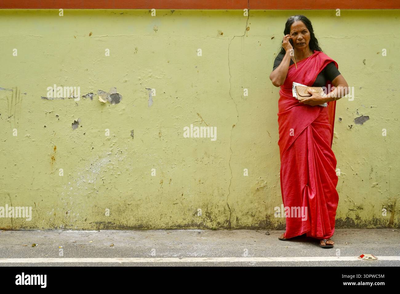 Ältere indische Frau in rotem Saree, die auf dem Handy spricht, steht an der bunten Wand in Alappuzha, Kerala, Indien Stockfoto