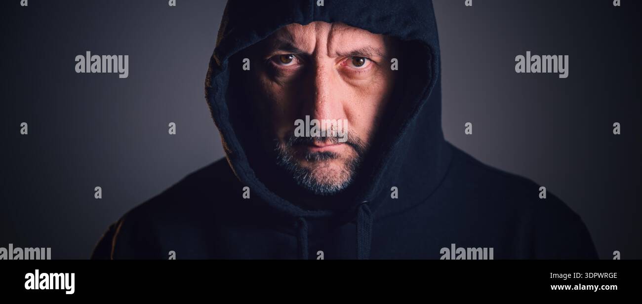 Porträt eines Mannes mittleren Alters mit kurzem Bart und verwittertem Gesicht, der einen dunklen Hoodie trägt und direkt mit gefurchten Augenbrauen und intensivem Ga starrt Stockfoto