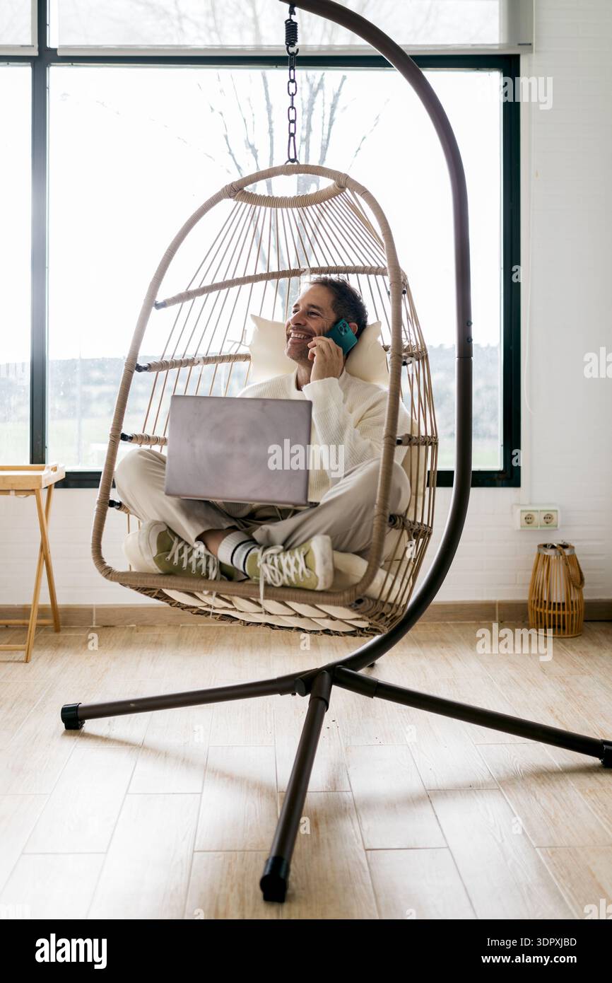 Lächelnder Mann, der von zu Hause aus arbeitet, während er im Kreuz in einem bequemen Hängesessel sitzt. Er spricht am Telefon und genießt einen Moment der Produktivität Stockfoto
