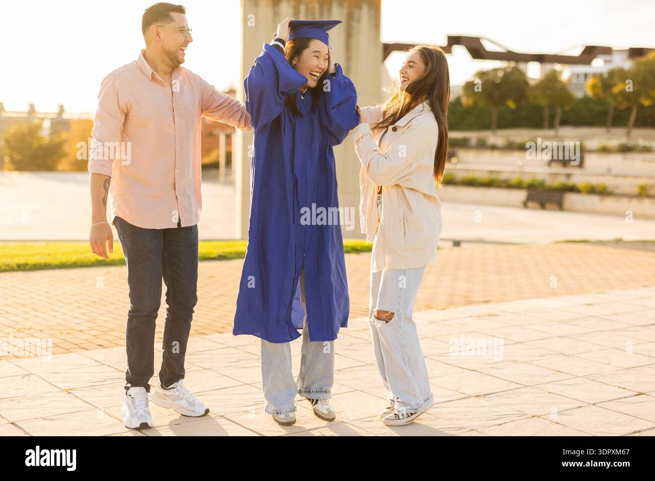 Drei Freunde feiern den Abschlussfeiertag im Freien. Der Absolvent trägt eine blaue Mütze und ein Kleid und wird von zwei Freunden fröhlich umarmt Stockfoto
