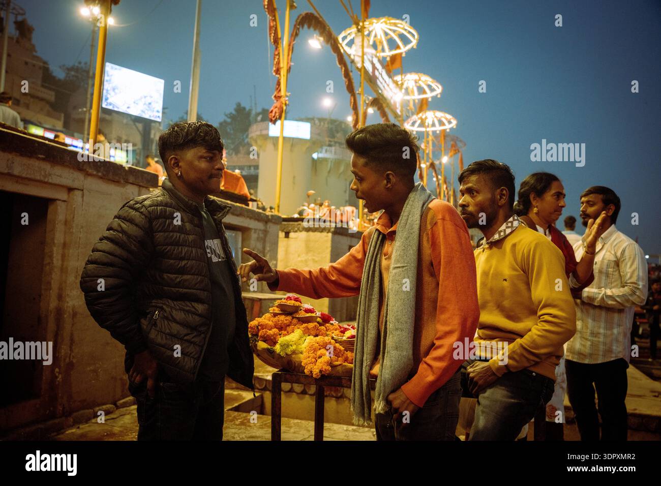 Männer sprechen in der Nähe von Blumenopfern auf einer Straße in Varanasi, Indien, während der Abendstunden mit festlichen Lichtern. Stockfoto